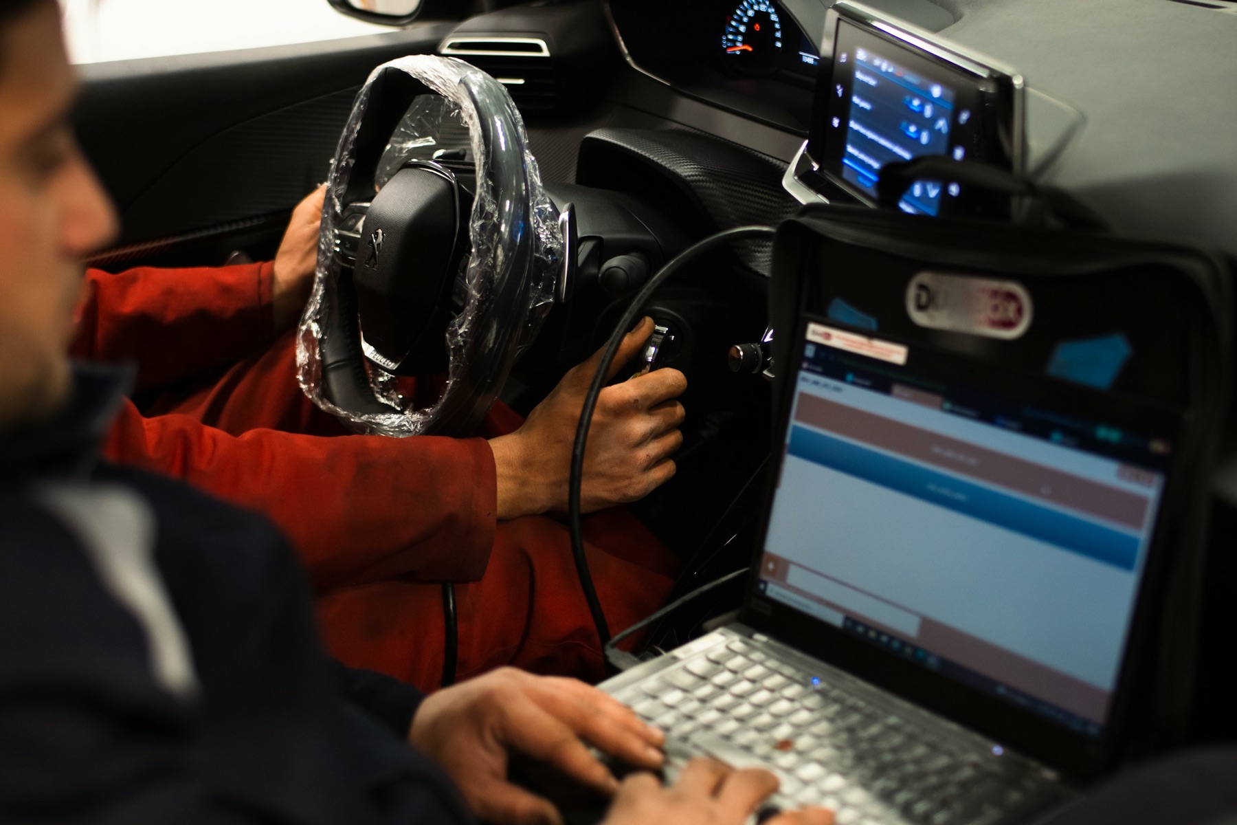 Technician using a diagnostic laptop connected to a vehicle in a workshop, with the steering wheel under a protective cover